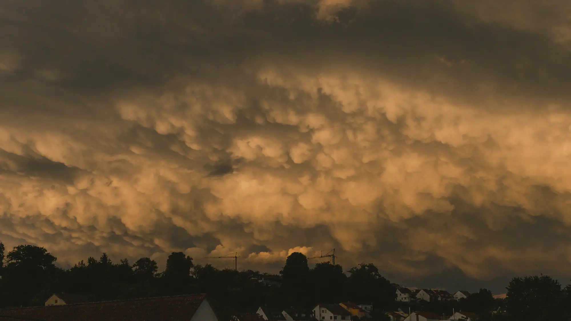 Low, pouched storm clouds lit amber at dusk over a line of trees and rooftops