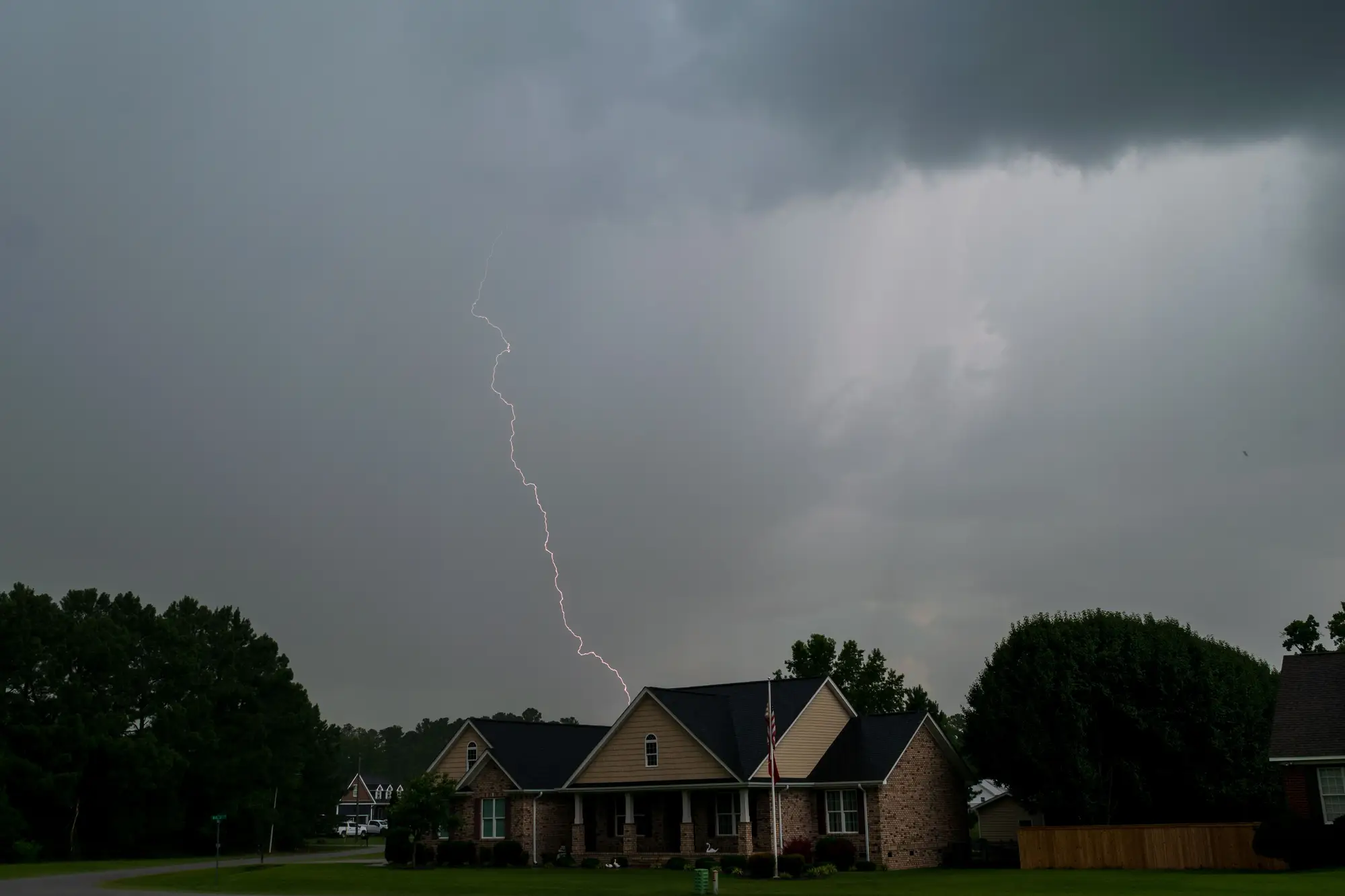 A thunderstorm with a lightning strike over a single-story brick house with an asphalt shingle roof